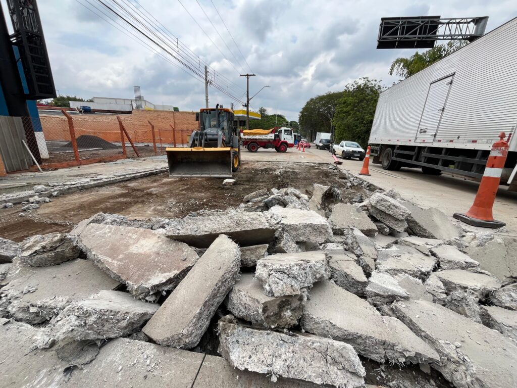 A imagem mostra uma cena de obra em uma rua, com grande quantidade de concreto quebrado espalhado no primeiro plano. Um trator com a pá levantada está posicionado ao lado do material quebrado, pronto para movê-lo. Ao fundo, um caminhão de carga com lona amarela está parado em meio ao tráfego, e outros veículos circulam na via. A cena sugere uma interrupção no trânsito devido aos trabalhos de pavimentação ou demolição. O céu está nublado, indicando condições climáticas instáveis.