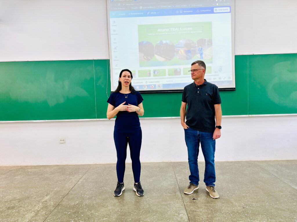 A imagem mostra uma sala de aula onde dois professores estão em pé em frente a um quadro verde e a um projetor. A mulher à esquerda veste uma blusa azul escura e calças combinando, enquanto o homem à direita usa uma camisa polo cinza e jeans. O projetor exibe uma apresentação com o título "Aluno TEA: Lucas" e várias imagens. O quadro verde está em branco, sugerindo que a aula está prestes a começar ou terminou. O chão da sala é de concreto e há um interruptor de luz na parede branca atrás dos professores.