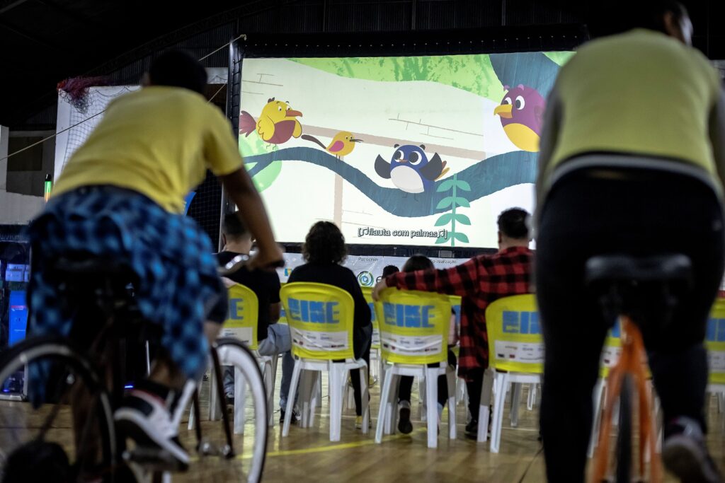 A imagem mostra um grupo de pessoas assistindo a uma projeção em uma tela grande de um desenho animado com pássaros. Há pessoas sentadas em cadeiras amarelas e outras em bicicletas, sugerindo uma exibição ao ar livre ou em um espaço amplo. A iluminação é baixa, com a tela sendo a principal fonte de luz. As cadeiras amarelas têm o logotipo "BIKE" estampado, indicando algum tipo de patrocínio ou evento relacionado a bicicletas. A tela exibe um cenário natural com árvores e vários pássaros animados em cores vibrantes. A cena transmite uma atmosfera de lazer e entretenimento comunitário.