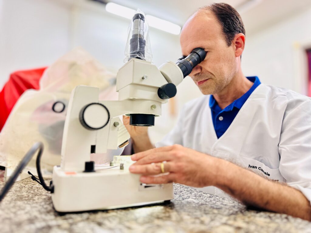 A imagem mostra um homem de meia-idade usando um jaleco branco de laboratório e um colarinho azul, examinando algo através de um microscópio. Ele tem uma expressão concentrada no rosto enquanto ajusta o foco. O microscópio é branco e parece ser um equipamento de laboratório padrão. Ao fundo, há objetos cobertos com panos, sugerindo um ambiente de laboratório ou pesquisa. Há uma inscrição "Ivan Canale" no jaleco do homem.