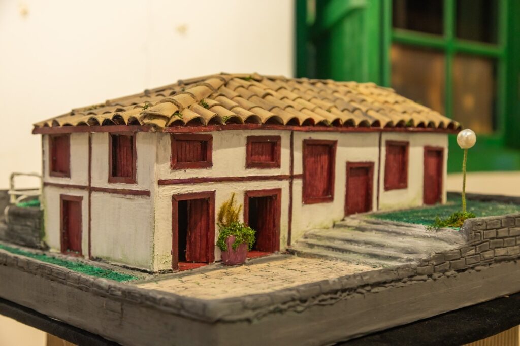 A miniatura de uma casa branca com detalhes em madeira escura e telhado de telhas marrons ocupa o centro da imagem. A casa possui duas portas frontais abertas, revelando o interior escuro, e várias janelas com persianas fechadas. A fachada é dividida horizontal e verticalmente por faixas de madeira escura. Um pequeno jardim com grama verde e um vaso com folhagem amarela fica em frente às portas. Uma escadaria de pedra cinza leva a um nível mais alto à direita da casa, onde há um pequeno mastro com uma esfera branca no topo, simulando um poste de luz. O cenário é completado por um muro de pedra cinza na base da estrutura e um fundo desfocado em tons de branco e verde.