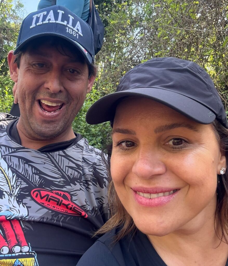 A imagem mostra um casal tirando uma selfie em um ambiente externo, com vegetação ao fundo. O homem, à esquerda, usa um boné azul escuro com a inscrição "ITALIA 1861" em branco e uma camiseta cinza com estampa de penas e um logo vermelho escrito "MAKI FISHING". Ele tem um sorriso largo e aberto, mostrando os dentes. A mulher, à direita, usa um boné preto e uma camiseta preta. Ela tem um sorriso gentil e usa um brinco de strass em sua orelha direita. A iluminação sugere que a foto foi tirada durante o dia, com luz natural.