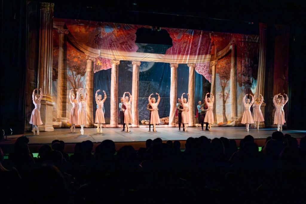 A imagem mostra uma cena de ballet em um palco, com bailarinos em movimento. No centro, um grupo de bailarinas vestidas com trajes cor-de-rosa claras realizam uma coreografia, com os braços erguidos em uníssono. Algumas delas estão em ponta. O cenário atrás delas é um painel pintado que retrata uma paisagem clássica, com colunas gregas, árvores e um céu noturno com uma lua proeminente. A iluminação do palco destaca os bailarinos, enquanto a plateia, na parte inferior da imagem, permanece na penumbra. Há também alguns bailarinos masculinos em trajes escuros, interagindo com as bailarinas em alguns pontos do palco.