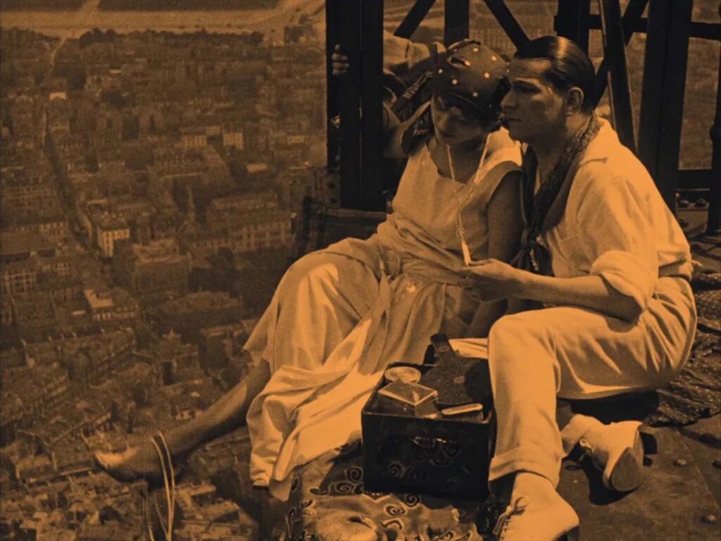A imagem mostra um casal sentado em uma plataforma alta, com uma cidade vista em perspectiva abaixo. O homem, à direita, veste uma camisa de manga comprida branca e calças claras, com um lenço colorido ao redor do pescoço. Ele tem o cabelo penteado para trás e olha para a mulher ao seu lado. A mulher, à esquerda, usa um vestido claro e um lenço estampado na cabeça. Ela está usando um colar de pérolas e também olha para algo que está segurando em suas mãos. Entre eles, há uma caixa ornamentada aberta, contendo diversos itens, possivelmente de maquiagem ou higiene pessoal, como espelhos e pincéis. A cena tem um tom sépia, característico de filmes antigos.