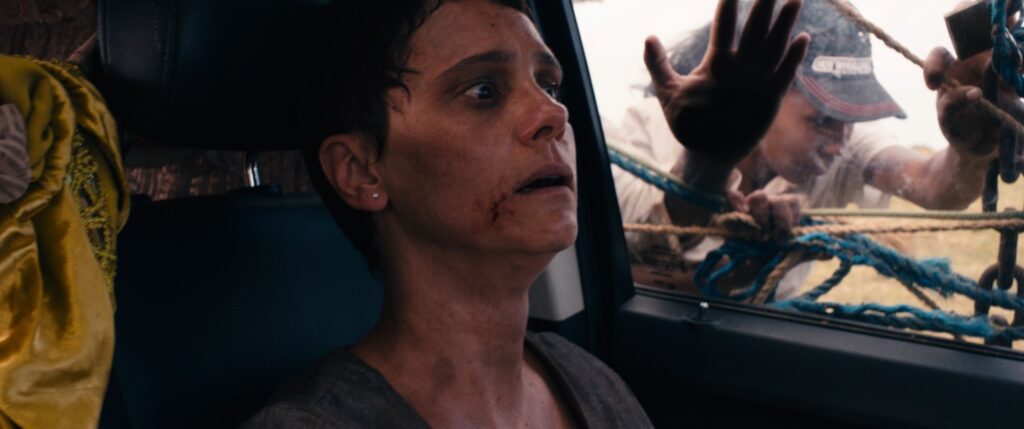 A imagem mostra uma cena tensa dentro de um veículo. Em primeiro plano, há uma mulher com expressão de medo ou choque, com os olhos arregalados e a boca ligeiramente aberta. Ela tem cortes e sangue no rosto, especialmente ao redor da boca e na bochecha, indicando que passou por alguma violência ou acidente. Ela veste uma camiseta cinza simples. Ao lado dela, parcialmente visível, está uma pessoa vestindo um tecido amarelo com detalhes dourados, possivelmente um traje tradicional ou cerimonial. Do lado de fora do veículo, através da janela, duas pessoas aparecem. Uma delas está estendendo a mão em direção ao veículo, vestindo um boné escuro, e a outra está ao fundo, perto de cordas grossas que parecem estar amarradas ou penduradas, criando uma barreira visual. A iluminação da cena é dramática, com sombras fortes, aumentando a sensação de perigo e tensão. O ambiente externo parece ser uma área aberta, talvez rural, e o conjunto da imagem sugere uma situação de ameaça iminente, como uma perseguição ou tentativa de captura. A perspectiva mostra o interior do carro, transmitindo a sensação de confinamento e vulnerabilidade da mulher.