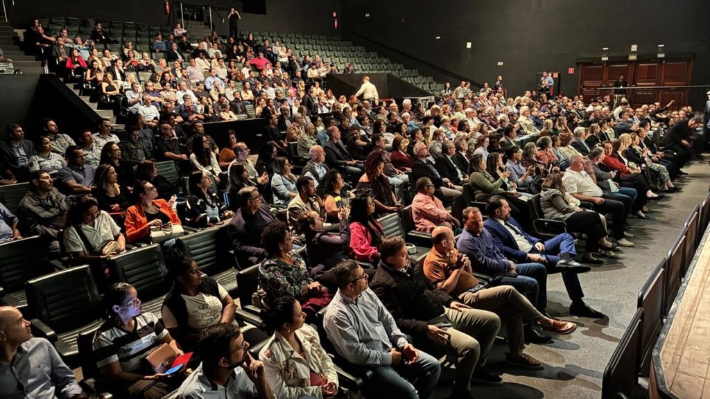 A imagem mostra uma plateia sentada em um teatro ou auditório, voltada para um palco que não está visível. As pessoas estão sentadas em fileiras de assentos, a maioria delas olhando na mesma direção, indicando que um evento está em andamento. A iluminação é baixa, típica de um ambiente de apresentação. A audiência é diversificada em termos de idade e vestimenta, com pessoas usando roupas casuais e outras mais formais. Alguns indivíduos estão segurando celulares ou cadernos, possivelmente para anotações ou para registrar o evento. A arquitetura do local sugere um espaço moderno, com assentos em tons escuros e paredes pretas ao fundo. O ambiente parece estar cheio, com poucas cadeiras vazias visíveis.