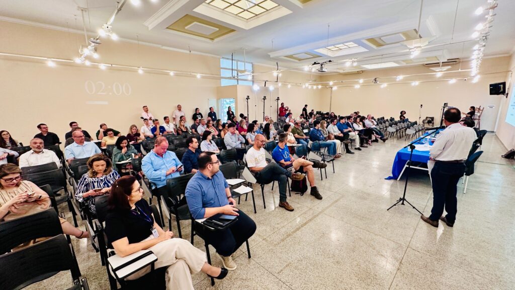 A imagem mostra uma sala de conferências cheia de pessoas assistindo a uma apresentação. A sala é bem iluminada, com luzes no teto que incluem um design central decorativo. Um grande relógio digital na parede indica 02:00. As pessoas estão sentadas em fileiras de cadeiras, voltadas para o palco, onde um apresentador está em pé atrás de uma mesa coberta por um pano azul e usando um microfone. O apresentador veste uma camisa clara e calças escuras, e está de costas para a câmera. A audiência é composta por homens e mulheres, vestidos de maneira casual e formal, alguns prestando atenção ao palestrante, outros olhando para seus celulares ou cadernos. O piso é de azulejos claros, refletindo a luz do ambiente. A atmosfera parece ser de concentração e formalidade, típica de uma palestra ou reunião profissional.