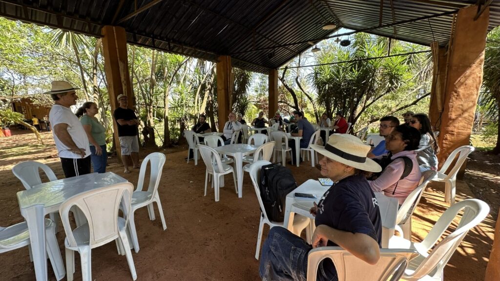 A imagem mostra um grupo de pessoas reunidas sob uma cobertura rústica, possivelmente um quiosque ou área de lazer ao ar livre. Há várias mesas e cadeiras de plástico brancas dispostas no local, com algumas pessoas sentadas e outras em pé. No primeiro plano, à esquerda, um homem com chapéu de palha branco e camiseta branca está conversando com uma mulher de camiseta verde, enquanto um homem de camiseta preta e óculos observa. Mais ao fundo, várias pessoas estão sentadas à mesa, algumas conversando entre si e outras parecendo escutar. O ambiente é cercado por vegetação exuberante, com árvores e palmeiras visíveis, indicando um local natural e possivelmente tropical. A cobertura é feita de telhas escuras, apoiada em pilares de madeira e alvenaria aparente. A luz do sol ilumina a cena, criando sombras interessantes no chão de terra batida.