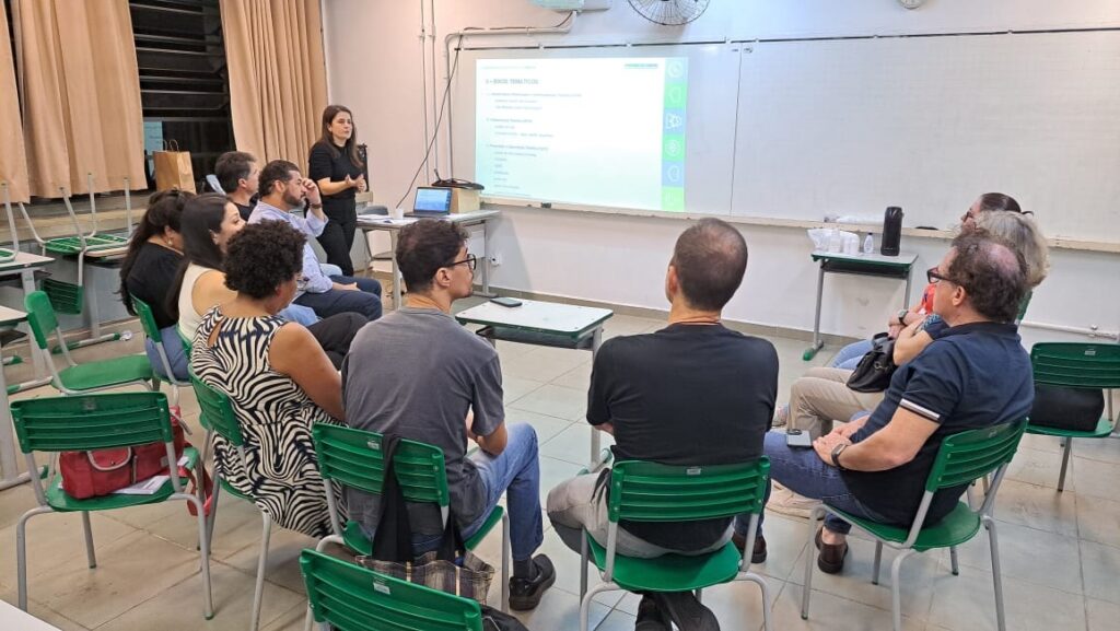 Uma mulher está em pé em frente a uma tela de projeção, apresentando algo para um grupo de pessoas sentadas em cadeiras em uma sala de aula. A tela exibe um slide com texto e ícones. As pessoas na plateia parecem estar prestando atenção à apresentação. A sala de aula tem carteiras e cadeiras verdes, e as cortinas nas janelas estão fechadas.