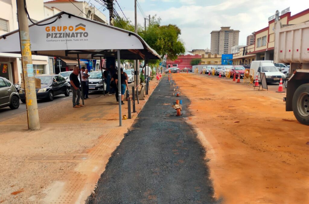 A imagem mostra uma rua em obras, com a calçada recém-pavimentada com asfalto escuro, contrastando com a terra alaranjada ao lado, que ainda não foi trabalhada. Há algumas pessoas na calçada, observando a situação, e vários carros estacionados. Na lateral da calçada, um toldo branco com a inscrição "GRUPO PIZZINATTO" oferece sombra. Mais adiante, a rua continua com a terra exposta, delimitada por cones de sinalização e algumas barreiras. Ao fundo, prédios comerciais e residenciais compõem o cenário urbano. Um caminhão de grande porte, provavelmente envolvido na obra, está estacionado à direita da imagem.