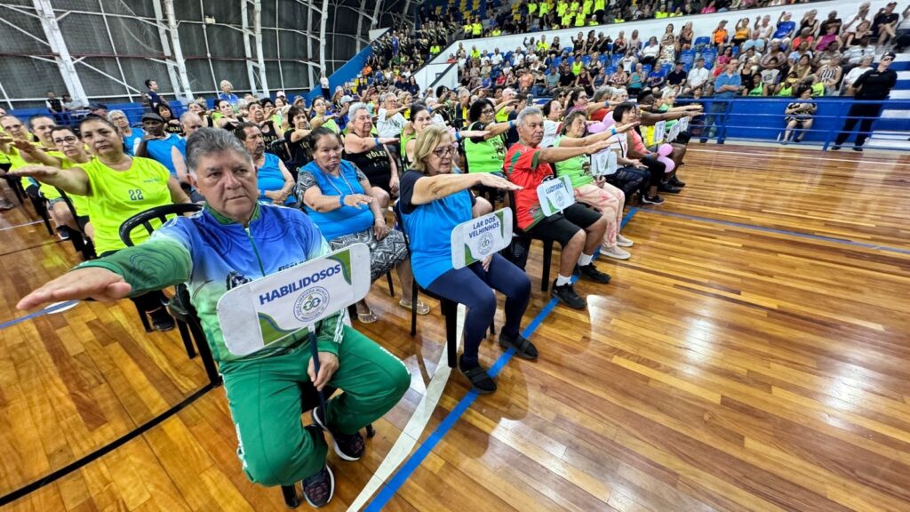 A imagem mostra um grupo de idosos sentados em cadeiras em uma quadra esportiva, todos realizando um exercício com os braços estendidos para a frente. Alguns dos participantes estão segurando placas com dizeres em português, como "HABILIDOSOS" e "LAR DOS VELHINHOS". Os idosos vestem roupas esportivas de cores variadas, predominando o verde e o azul. Ao fundo, outras pessoas estão sentadas em arquibancadas, assistindo à atividade. O piso da quadra é de madeira, com marcações azuis e brancas. A iluminação é artificial, típica de ambientes fechados. A atmosfera parece ser de um evento esportivo ou de atividade física voltada para a terceira idade, com um clima de participação e engajamento.