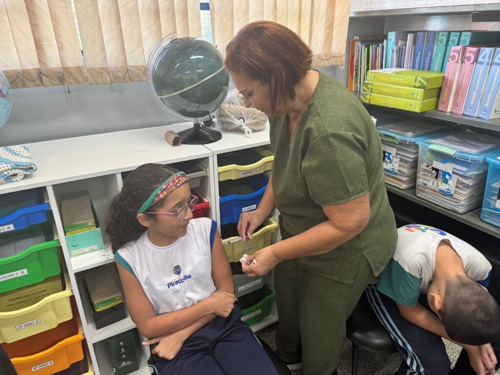 A professora, vestida com um uniforme verde escuro, está em pé e se inclina para uma menina sentada, que usa óculos e uma tiara colorida. A professora parece estar entregando algo para a menina, que olha para ela com atenção. Ao fundo, há uma estante cheia de livros e materiais escolares, incluindo um globo terrestre e caixas organizadoras coloridas. Um menino, de camiseta branca e azul, está curvado sobre o colo da professora, com a cabeça baixa. A cena sugere um ambiente escolar, possivelmente uma sala de aula ou biblioteca, onde a professora está interagindo com os alunos.
