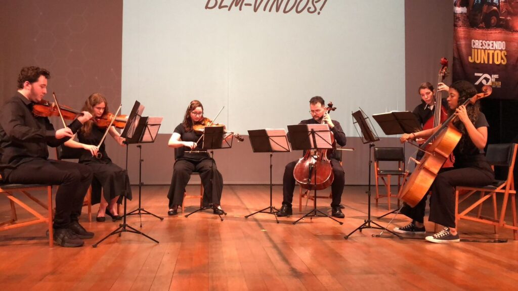 A imagem mostra um grupo de músicos se apresentando em um palco. Há seis músicos no total, todos vestidos com roupas pretas. Eles estão tocando violinos e violoncelos. O palco tem um piso de madeira e um fundo branco com algumas projeções. À direita, há um banner com a inscrição "CRESCENDO JUNTOS". Os músicos estão sentados em cadeiras e cada um tem um atril com partituras. A iluminação do palco é suave, criando uma atmosfera profissional e elegante.