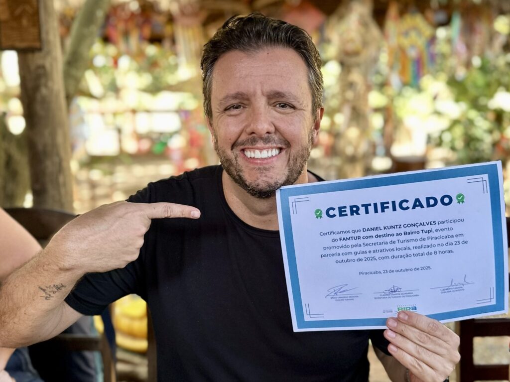 O homem sorri para a câmera, segurando um certificado e apontando para ele com o dedo indicador. O certificado, com bordas azuis e brancas, declara que "DANIEL KUNTZ GONÇALVES participou do FAMTUR com destino ao Bairro Tupi". O evento foi promovido pela Secretaria de Turismo de Piracicaba em parceria com guias e atrações locais, e ocorreu em 23 de outubro de 2025, com duração total de 8 horas. O local e a data de emissão do certificado são Piracicaba, 23 de outubro de 2025. Abaixo, há assinaturas de Diego Camargo Medeira (Guia de Turismo), Cleverson Guariaria (Secretaria de Turismo de Piracicaba) e Roberta Carajol (Chácara Catamento), juntamente com o logotipo "PIRA". O homem está vestindo uma camiseta preta e tem uma tatuagem em seu braço esquerdo. O fundo da imagem está desfocado, mostrando elementos que parecem ser de um ambiente externo com vegetação e decorações coloridas.