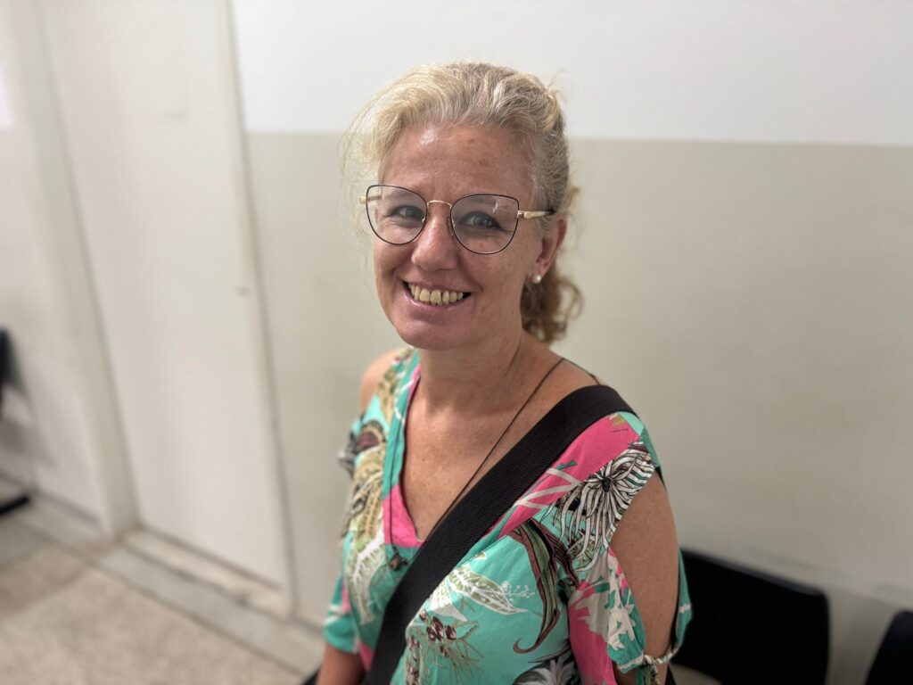 A mulher sorri para a câmera, usando óculos de aro dourado e uma blusa de alças finas com estampa floral colorida. Seu cabelo loiro está preso em um coque desfeito, com algumas mechas soltas emoldurando seu rosto. Ela usa brincos de pérola e um colar fino com um pingente escuro. A luz ilumina seu rosto, destacando suas sardas e a textura de sua pele. Ao fundo, uma parede branca e um corredor com pouca iluminação criam um ambiente simples e neutro.