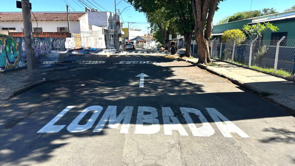 A imagem mostra uma rua residencial em um dia ensolarado. Na estrada, há uma faixa pintada de branco com a palavra "LOMBADA" escrita nela, indicando uma lombada ou redutor de velocidade. Acima da palavra "LOMBADA", há uma seta branca pintada no asfalto, apontando para a frente, indicando a direção do tráfego. Mais adiante na estrada, há outra inscrição em branco que diz "ESCOLA", com um sinal de trânsito com um "E" dentro de um círculo vermelho ao lado. À esquerda da estrada, há um muro de tijolos coberto de grafites coloridos. Uma parede branca com janelas e um telhado de telhas vermelhas se ergue atrás do muro. Há também um poste de luz com cabos elétricos suspensos acima. À direita da estrada, há uma calçada com grama crescendo ao lado. Uma cerca de arame com postes de concreto delimita a propriedade. Atrás da cerca, há um edifício verde com janelas escuras e um telhado de metal. Árvores altas com folhagem densa projetam sombras sobre a estrada e a calçada. O céu está limpo e azul.