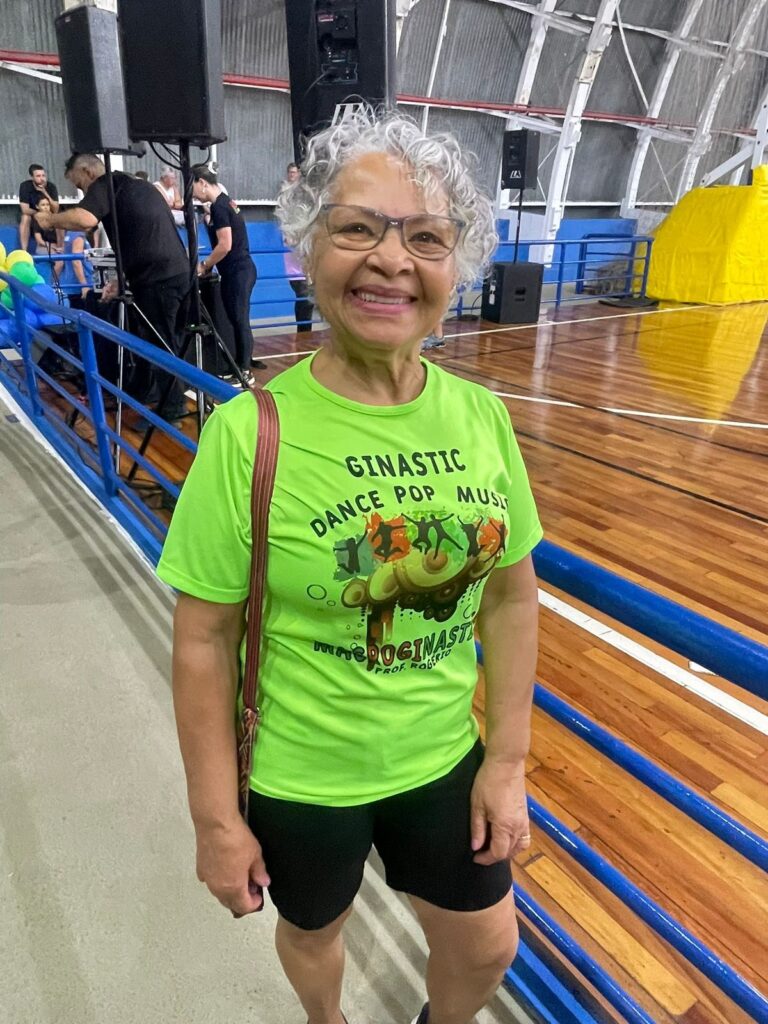 A mulher sorri para a câmera, vestindo uma camiseta verde neon com a estampa "Ginacstic Dance Pop Music Prof. Roberto". Ela usa óculos e tem cabelo grisalho cacheado. Ela está em uma quadra poliesportiva com piso de madeira, cercada por grades azuis. Ao fundo, há equipamentos de som, pessoas e uma estrutura amarela.