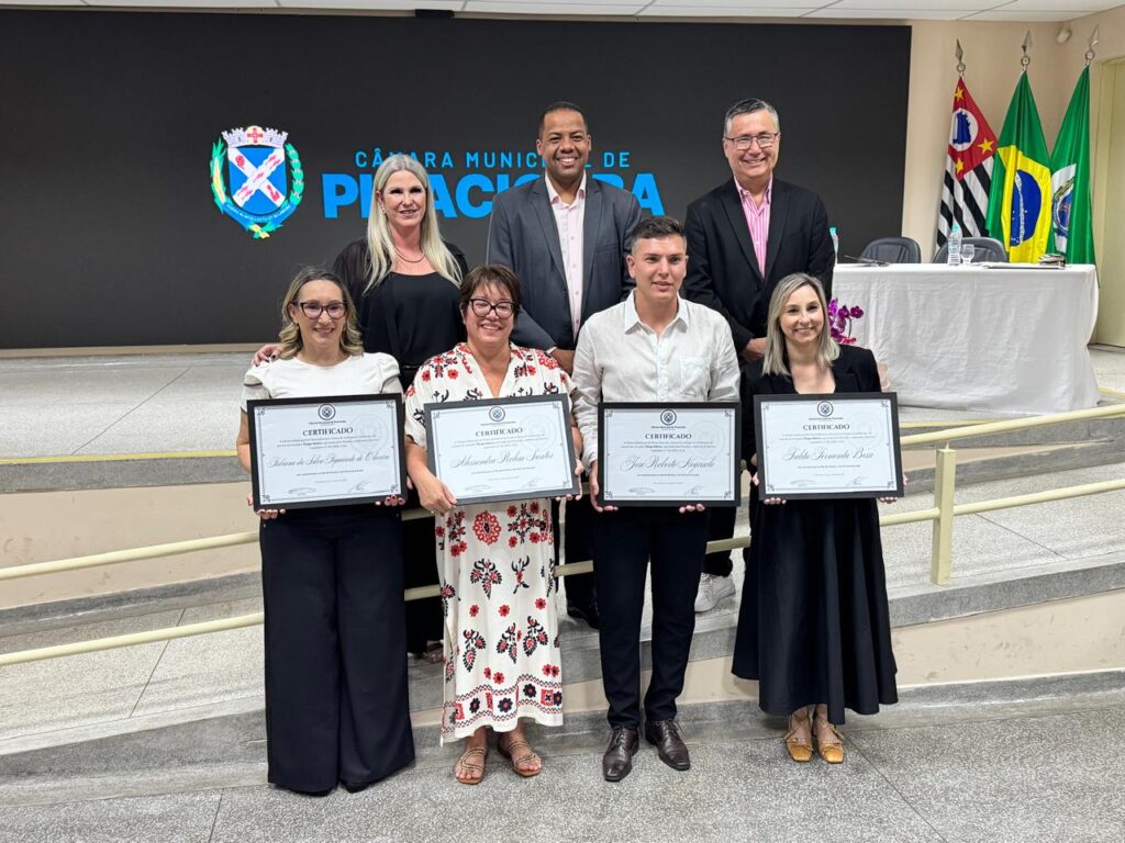 Oito pessoas estão posando para uma foto em um ambiente formal, provavelmente durante um evento da Câmara Municipal de Piracicaba. Quatro delas, na primeira fila, seguram certificados em molduras, indicando que foram homenageadas ou reconhecidas. Ao fundo, há uma tela grande com o nome “Câmara Municipal de Piracicaba” e bandeiras do Brasil e do estado de São Paulo, reforçando o caráter oficial da ocasião. Todos estão vestidos com roupas sociais, e o clima é de celebração e reconhecimento público.