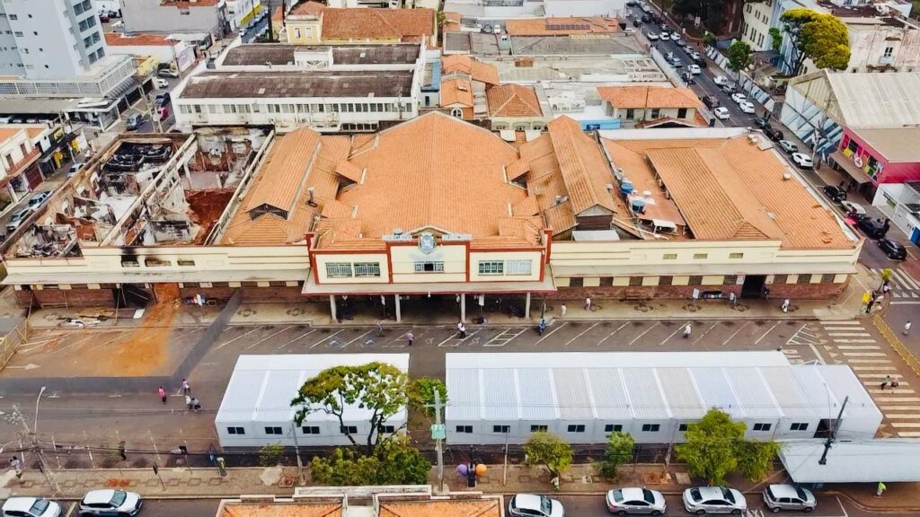 A imagem, tirada de um ângulo elevado, mostra uma vista aérea de uma área urbana com um edifício proeminente que parece ter sofrido danos significativos, possivelmente devido a um incêndio. O edifício principal, com telhado de telhas laranjas, exibe uma seção central onde o telhado e as paredes internas foram destruídos, expondo a estrutura interna e detritos. Ao redor desta área danificada, o resto do edifício parece intacto, com fachadas em tons de creme e detalhes em tijolo. Há uma entrada central com um brasão e janelas em cada lado. Em frente ao edifício principal, uma rua com marcações de estacionamento está parcialmente ocupada por duas grandes estruturas temporárias, semelhantes a tendas ou barracões modulares, com telhados brancos e janelas retangulares. Pessoas e alguns carros são visíveis na área, sugerindo atividade contínua apesar dos danos. Ao redor do edifício principal e das estruturas temporárias, outros edifícios da cidade podem ser vistos, alguns mais altos e modernos, outros com telhados de telha semelhantes ao edifício danificado. Carros estão estacionados e circulando nas ruas adjacentes, indicando que a vida urbana continua ao redor da área afetada. A vegetação, incluindo árvores, também está presente, adicionando um toque de cor à cena urbana.