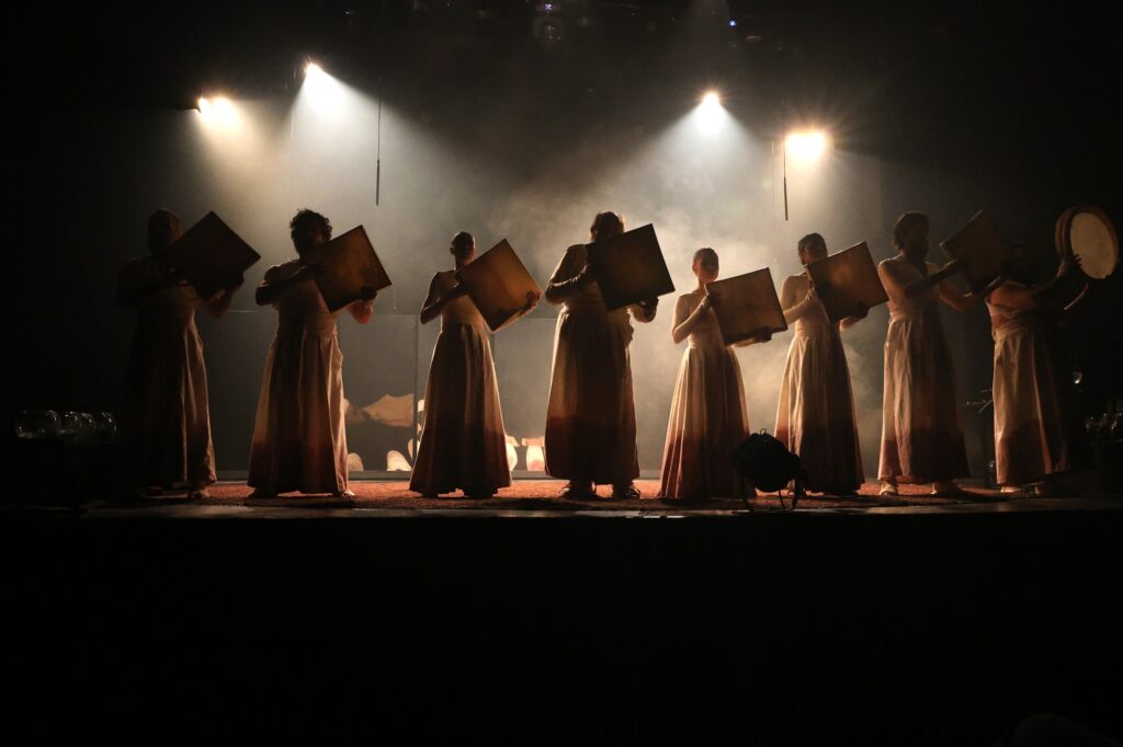 A imagem mostra um grupo de pessoas em um palco escuro, iluminadas por fortes focos de luz vindos de cima. Todos estão vestidos com longas vestes claras, e a maioria segura um objeto quadrado e plano em frente ao corpo. Um dos indivíduos na extremidade direita segura um tambor redondo. A iluminação cria silhuetas distintas, com a fumaça ou névoa no palco realçando os feixes de luz. A atmosfera é dramática e performática.