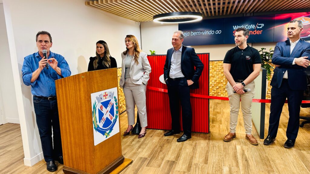 A imagem mostra um evento de inauguração, possivelmente de um espaço de coworking ou café, com a presença de várias pessoas em traje formal e informal. No centro da imagem, um homem de camisa azul e calça jeans está em pé atrás de um púlpito de madeira, segurando um microfone e falando. O púlpito possui um brasão com o lema "AUDAX IN INTELLECTU ET IN LAI". À direita do homem no púlpito, há três homens e uma mulher. Uma mulher de blazer cinza e calça social está com as mãos nos bolsos. Ao lado dela, um homem de terno escuro sorri, com as mãos nos bolsos. Mais à direita, um homem com uma camiseta preta com o logo "Work Café Santander" e calça bege segura um microfone. Por fim, o homem mais à direita, vestindo um terno azul escuro, está de braços cruzados. Ao fundo, uma parede com um painel de madeira e um letreiro luminoso que diz "Seja bem-vindo ao Work Café Santander". Uma fita vermelha atravessa a cena, indicando uma área de inauguração. O piso é de madeira clara, com um padrão que lembra tábuas. A iluminação é clara e moderna, com um anel de luz circular no teto.