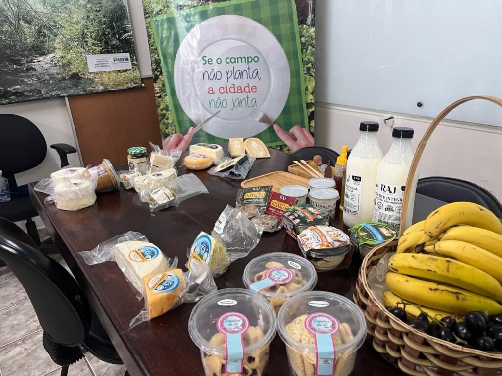 A imagem mostra uma mesa de madeira repleta de produtos alimentícios, com foco em queijos e laticínios. Há uma variedade de queijos embalados individualmente, alguns em formatos redondos e outros em cunhas. Também há potes de iogurte ou creme de queijo, garrafas de leite e um pequeno pote de geleia. No canto inferior direito, há uma cesta de vime contendo cachos de bananas e uvas escuras. Na mesa, também se observam pacotes de biscoitos em embalagens transparentes e alguns temperos ou condimentos. Ao fundo, há um cartaz com a frase "Se o campo não planta, a cidade não janta", com ilustrações de um prato e mãos segurando um garfo. Um outro cartaz com o logo "PIRA" e imagens de paisagens naturais também está visível. Há também cadeiras de escritório ao redor da mesa. A iluminação sugere um ambiente interno, possivelmente um escritório ou sala de reuniões.