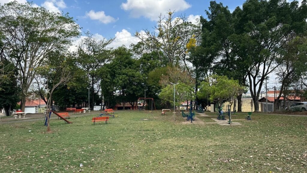 A imagem mostra um parque com gramado verde e árvores frondosas, sob um céu azul com nuvens brancas. Há equipamentos de ginástica ao ar livre e um parquinho infantil com balanços e um escorregador. Bancos de madeira laranja estão espalhados pelo parque, convidando para um descanso. Ao fundo, casas com telhados vermelhos e um carro branco estacionado complementam a cena. A iluminação sugere um dia ensolarado, com sombras projetadas no chão.