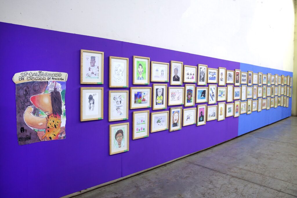 A imagem exibe uma exposição de arte em uma parede roxa vibrante. Várias obras de arte, a maioria delas retratos e desenhos animados emoldurados em madeira clara, estão penduradas em fileiras. À esquerda, um cartaz grande e colorido, enrolado na parte superior, anuncia "11º SALÃOZINHO DE HUMOR DE PIRACICABA". A arte no cartaz é um desenho caricatural com cores fortes. As obras penduradas na parede variam em estilo, com alguns retratos realistas e outros desenhos mais estilizados e cômicos. As molduras são uniformes, criando uma sensação de ordem na exibição. A iluminação destaca as obras e a cor da parede, criando um ambiente de galeria. A parede muda de cor para um tom mais claro de azul em uma seção mais distante, onde mais quadros continuam em exibição. O chão é de concreto aparente.