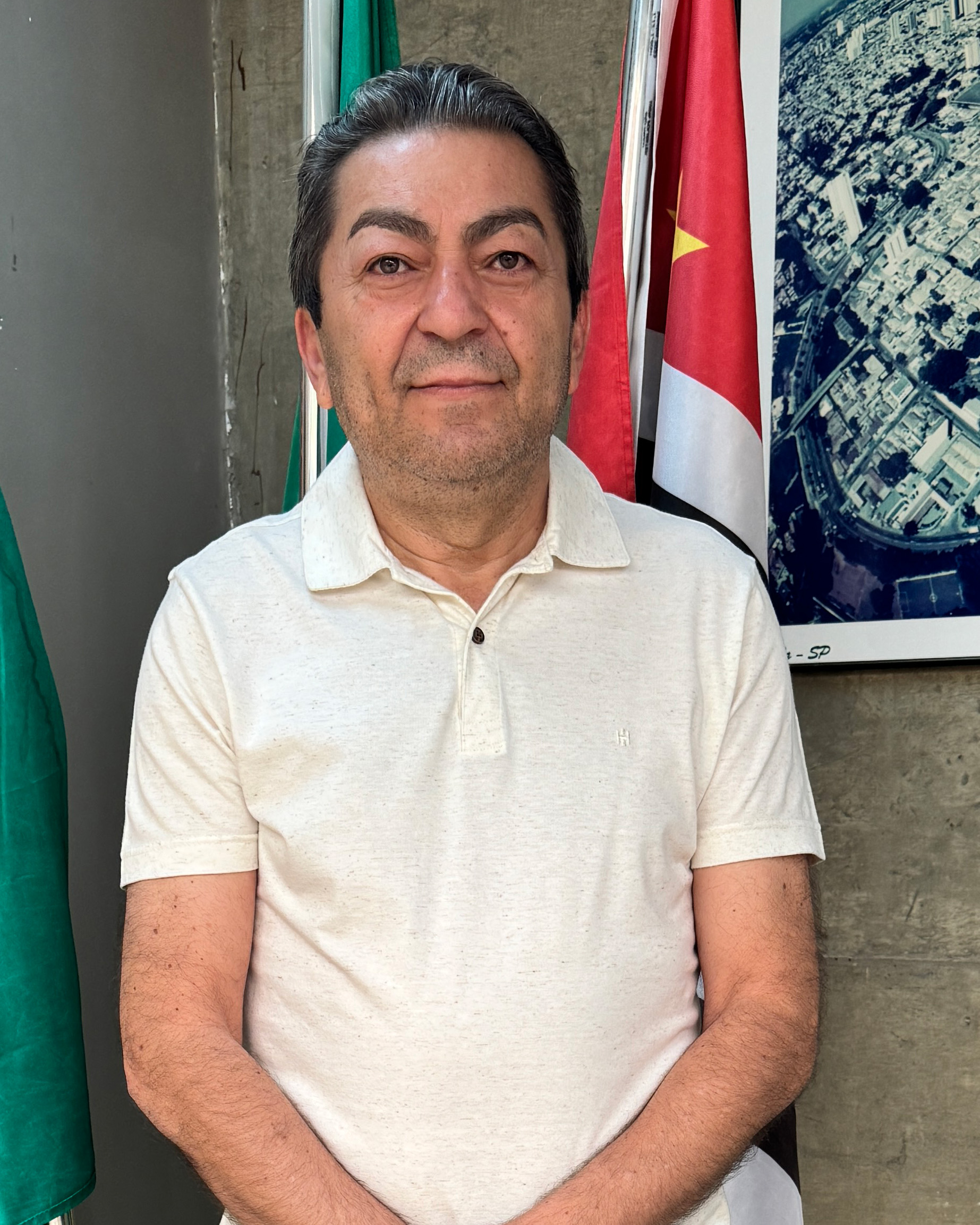 O homem na foto tem pele clara, cabelo escuro com mechas grisalhas, e usa uma camisa polo bege clara com um pequeno logo "H" bordado no peito esquerdo. Ele tem uma barba rala e um sorriso leve. Atrás dele, há duas bandeiras, uma verde e outra vermelha com uma estrela amarela, e uma foto aérea de uma cidade.