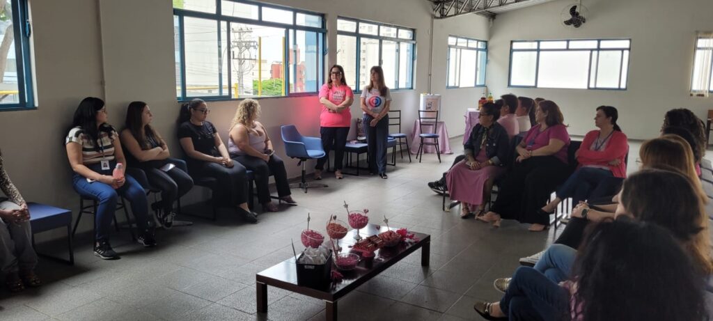 A imagem mostra um grupo de pessoas, a maioria mulheres, reunidas em uma sala. Algumas mulheres estão sentadas em cadeiras dispostas em semicírculo, enquanto outras estão em pé. Duas mulheres em pé, na parte frontal da sala, parecem estar liderando ou apresentando algo para o grupo. Uma delas veste uma camiseta rosa com um texto escrito, e a outra veste uma camiseta branca com um círculo azul e vermelho. Há uma mesa baixa à frente do grupo sentado, com recipientes de vidro contendo doces coloridos, possivelmente para um evento ou celebração. As janelas da sala são grandes e permitem a entrada de luz natural, revelando um pouco do ambiente externo com prédios e vegetação. O teto tem um ventilador de teto e uma estrutura metálica aparente. A atmosfera geral sugere uma reunião, palestra ou evento informal.