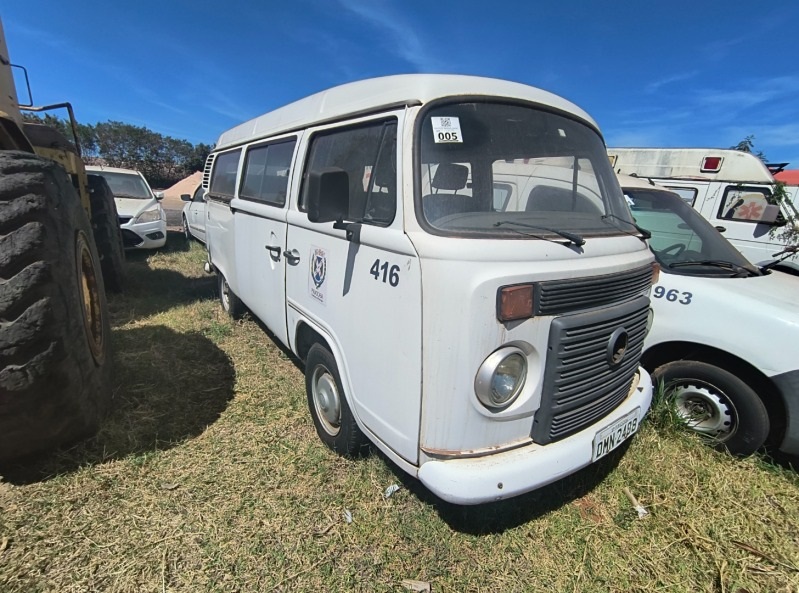 A imagem exibe uma van branca, possivelmente um modelo Volkswagen Kombi, estacionada em um terreno gramado sob um céu azul claro. A van parece estar em um pátio de veículos, com outros carros e um trator visíveis ao fundo e nas laterais. Detalhes notáveis incluem um adesivo com o número "005" no para-brisa, o número "416" pintado na lateral da porta e um distintivo com um brasão que parece ser de alguma instituição. A placa do veículo, parcialmente visível, é "DMN 2499". Ao lado da van, um carro branco com o número "963" pintado na porta e parte de um trator com um pneu grande e amarelo completam a cena. Ao fundo, é possível ver o que parecem ser ambulâncias. A vegetação ao redor é rasteira e seca, indicando um ambiente externo e possivelmente um local de descarte ou leilão de veículos.