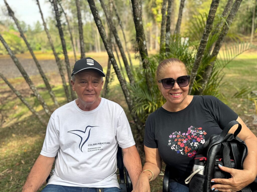 A imagem mostra um casal sentado em um ambiente externo, possivelmente um parque ou jardim, com árvores e folhagens ao fundo. O homem, à esquerda, veste uma camiseta branca com um desenho de beija-flor e a inscrição "COLIBRI PIRACICABA PARKINSON COM QUALIDADE DE VIDA", além de um boné preto. Ele parece estar em uma cadeira de rodas. A mulher, à direita, usa uma camiseta preta com um coração formado por flores coloridas e óculos escuros. Ela também parece estar em uma cadeira de rodas, e segura uma mochila preta com uma garrafa de água. Ambos sorriem para a câmera, transmitindo uma expressão de contentamento. A luz natural ilumina a cena, criando um ambiente agradável e sereno.