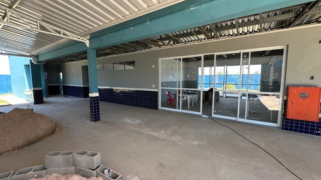 A imagem mostra o interior de um espaço em construção, possivelmente um restaurante ou área de convivência, com vista para o mar. O piso é de concreto bruto, e há uma grande pilha de areia em primeiro plano, juntamente com blocos de concreto empilhados. As paredes internas são pintadas de cinza, com uma faixa inferior revestida com azulejos azuis escuros. Colunas azul-petróleo sustentam o teto, que é exposto, com vigas metálicas e fiação aparente. Uma grande parede de vidro com caixilhos brancos se estende pela lateral direita, oferecendo uma vista clara do exterior, onde se pode ver o mar azul e a vegetação. Dentro desse espaço envidraçado, há uma mesa de trabalho e uma cadeira vermelha. No canto direito, há uma caixa de hidrante laranja, também com azulejos azuis escuros ao redor. Ao fundo, à esquerda, uma parede azul se destaca, com uma área gramada visível do lado de fora. A iluminação parece ser natural, vinda da área envidraçada e de outras aberturas.