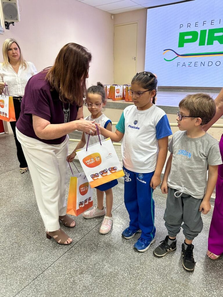 Uma mulher de cabelos castanhos, vestindo uma blusa roxa e calças brancas, entrega sacolas de presente para duas crianças. Uma das crianças, uma menina de óculos e cabelo preso, está de pé ao lado de outra menina de óculos. Atrás delas, um menino de óculos e camiseta cinza com a palavra "JORDAN" escrita nela está de pé. Ao fundo, um telão exibe o logotipo "PREFEITURA PIRACICABA FAZENDO MAIS". Há outras sacolas de presente dispostas em uma mesa ao fundo. A cena parece ser um evento escolar ou comunitário.
