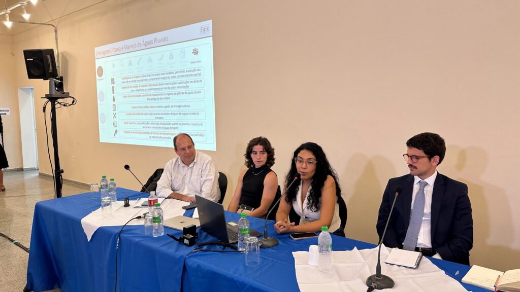 A imagem mostra uma apresentação em um ambiente que parece ser uma sala de reunião ou conferência. Na parede, há um projetor exibindo um slide com o título "Drenagem Urbana e Manejo de Águas Pluviais". O slide está dividido em duas seções principais: "Diagnóstico" e "Análise Crítica". Embaixo dessas seções, há uma lista de ações e recomendações relacionadas à gestão da drenagem urbana, como atualização do cronograma de obras, implantação de medidas de controle institucional, mapeamento das galerias de águas pluviais, cadastro técnico, levantamento sobre lançamento clandestino de esgoto e programa de manutenção do sistema de macrodrenagem. À frente do projetor, há uma mesa coberta com toalhas azul e branca, onde estão sentadas quatro pessoas (três mulheres e um homem). Elas parecem estar participando da apresentação, com alguns equipamentos como microfones, garrafas de água, copos, notebooks e cadernos sobre a mesa. A atmosfera sugere um encontro técnico ou uma reunião de trabalho sobre o tema da drenagem urbana.