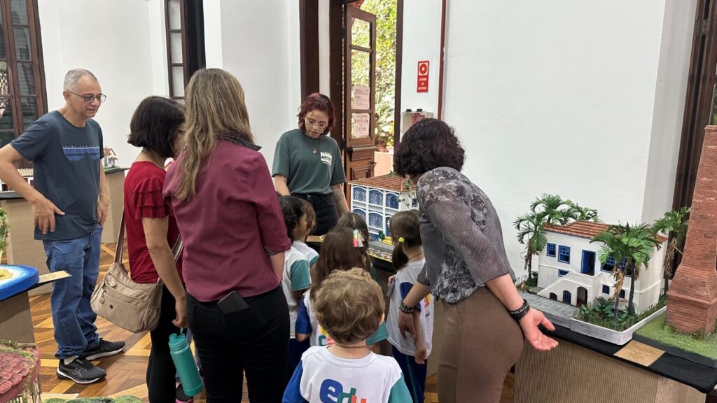 A imagem mostra um grupo de pessoas, incluindo crianças e adultos, observando miniaturas de construções em uma exposição. Uma mulher, possivelmente uma guia ou professora, está explicando algo às crianças, que estão visivelmente interessadas. Ao fundo, há uma porta aberta para um ambiente externo com vegetação, e nas paredes, quadros e um extintor de incêndio. As miniaturas parecem representar casas e edifícios históricos, com detalhes arquitetônicos e paisagismo em miniatura. O ambiente sugere um museu ou um espaço cultural educativo.
