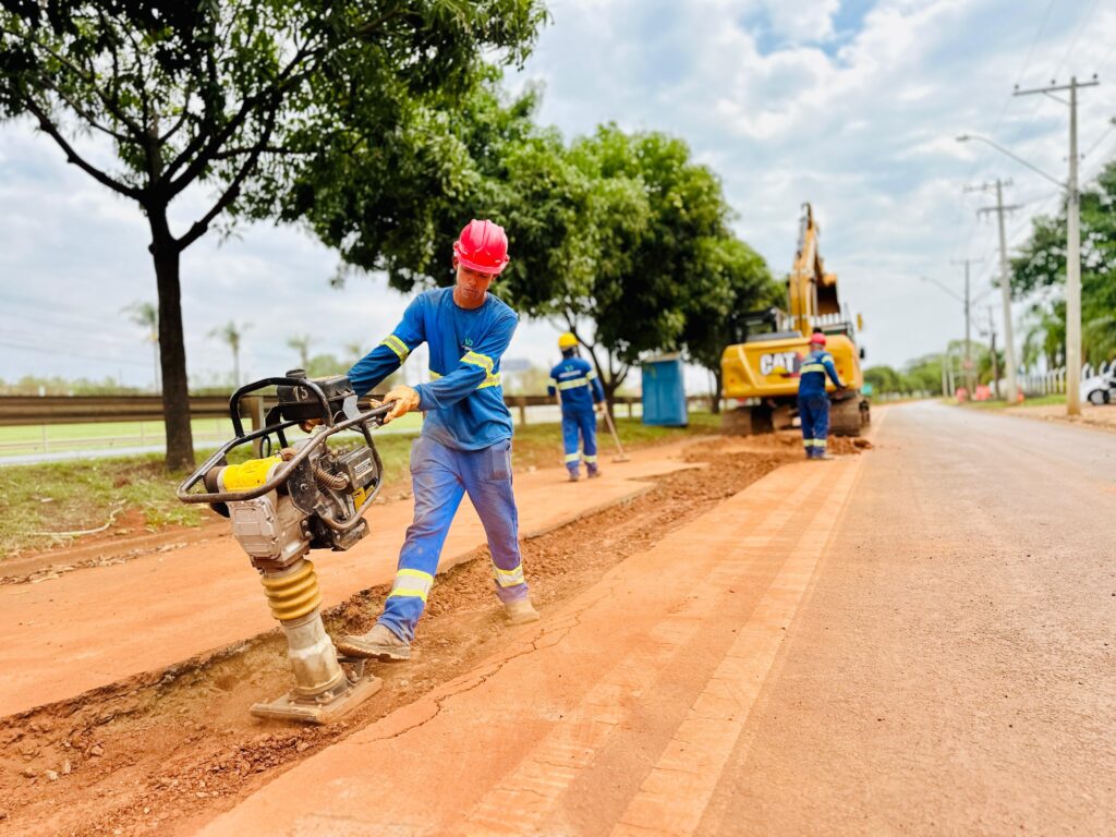 A imagem mostra uma equipe de trabalhadores em uma obra de construção de estrada. No primeiro plano, um trabalhador, vestindo uniforme azul com faixas refletivas amarelas e um capacete vermelho, está operando um compactador de solo. O equipamento está sendo usado para compactar o solo em uma vala recém-escavada ao lado da estrada. Ao fundo, outros dois trabalhadores estão presentes. Um deles, usando uniforme azul e capacete amarelo, parece estar varrendo o solo com uma vassoura. O outro, com uniforme azul e capacete vermelho, está perto de uma escavadeira amarela da marca CAT. A escavadeira está posicionada na lateral da estrada, com um pouco de terra ao redor de suas esteiras. O ambiente é externo, com árvores verdes e um céu nublado ao fundo. Uma cabine sanitária azul também é visível entre as árvores. A estrada em construção parece ser de terra batida, com a área de trabalho demarcada.