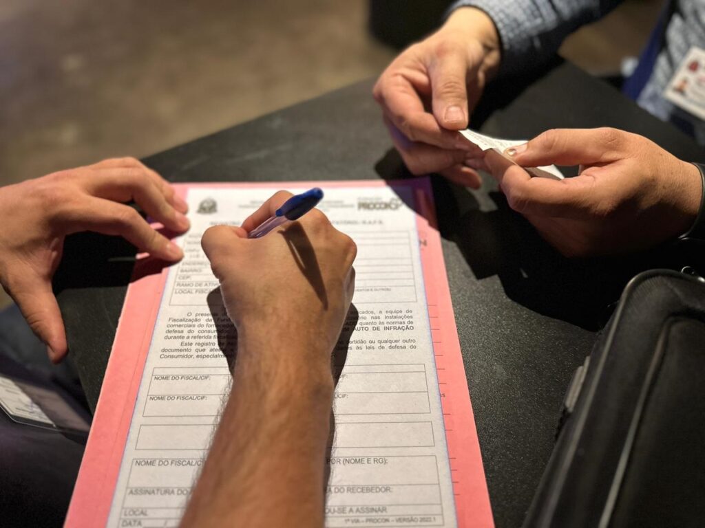 A imagem mostra um close-up de duas pessoas em uma mesa escura, preenchendo um formulário rosa. Uma pessoa está escrevendo no formulário com uma caneta azul, enquanto a outra pessoa segura um pequeno pedaço de papel. O formulário parece ser um documento oficial, com campos para "NOME DO FISCAL/CIF", "RAMO DE ATIV", "LOCAL FISC", "ASSINATURA DO", "DATA", "NOME DO RECEBEDOR", entre outros. O texto no formulário inclui frases como "O presente registro atesta...", "Fiscalização da Fund...", "defesa do consumidor" e "AUTO DE INFRAÇÃO", sugerindo que se trata de um documento relacionado a uma fiscalização ou auto de infração. O logotipo "PROCON" é visível no topo do formulário. A iluminação foca nas mãos e no formulário, com o fundo desfocado.