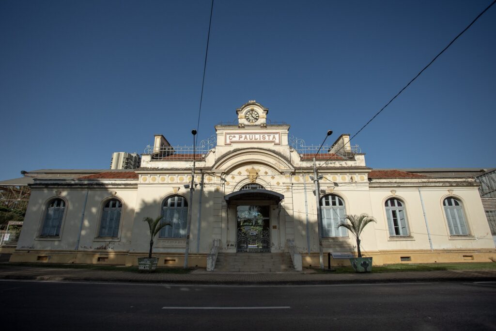 A imagem mostra a fachada de um edifício histórico com um telhado de telhas vermelhas e paredes de cor creme. O edifício tem uma arquitetura clássica, com janelas em arco e detalhes ornamentais. No centro da fachada, há uma entrada principal com uma escadaria e uma porta de vidro com grades. Acima da entrada, há um relógio e uma placa com a inscrição "Cª PAULISTA". Duas palmeiras em vasos estão posicionadas em frente ao edifício, e a rua com asfalto e faixas brancas pode ser vista na parte inferior da imagem. O céu azul e limpo domina a parte superior da imagem, com alguns fios elétricos cruzando o céu.