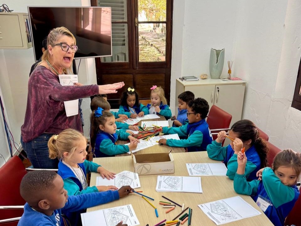 A professora, com óculos e um colar, está em pé e gesticula com a mão direita enquanto segura um papel. Ela está vestida com uma blusa vinho e jeans. À sua frente, sentadas em uma mesa de madeira, há várias crianças em idade escolar, vestindo uniformes azuis e verdes. Elas estão absortas em atividades de colorir, com lápis de cor espalhados pela mesa. Ao fundo, observa-se uma televisão grande na parede, uma estante com alguns objetos decorativos, como um vaso e porta-incenso, e uma janela com vista para uma área externa. A sala parece ser um ambiente de aprendizado, possivelmente uma sala de aula.