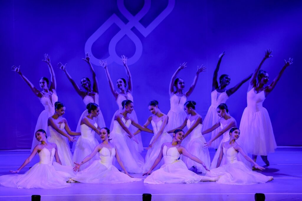 A imagem mostra um grupo de bailarinas em um palco, todas trajando vestidos brancos esvoaçantes. Elas estão sob uma iluminação azul vibrante, que cria uma atmosfera etérea e dramática. A maioria das bailarinas está em posições de dança clássica, com os braços erguidos e expressivos, enquanto outras estão em posições mais baixas, como se estivessem em pleno movimento. Ao fundo, um grande símbolo branco, que se assemelha a um nó ou a duas formas entrelaçadas, está projetado ou pintado no cenário, adicionando um elemento visual intrigante à cena. A composição geral sugere uma performance de balé, capturando a graça, a técnica e a beleza do movimento.