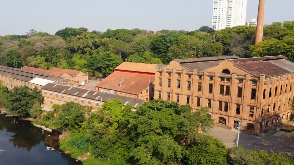 A imagem mostra um complexo de edifícios industriais antigos, feitos de tijolos aparentes, com telhados de telha aparente. Há uma grande chaminé de tijolos ao lado de um dos edifícios. Ao fundo, uma floresta densa e uma torre branca moderna, contrastando com as construções antigas. Um rio com águas escuras corre na frente dos edifícios, cercado por vegetação exuberante.