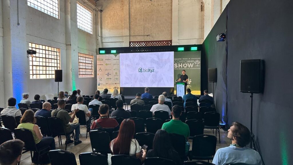 A imagem mostra um evento em um grande salão com arquitetura industrial. Há um palco com um telão onde se lê "BioXyz" e "Encontro Técnico de Compostagem Compo 2025 Show". Um homem está em pé no palco, apresentando. Ao redor, uma audiência está sentada em cadeiras, assistindo à apresentação. As paredes do salão são altas e claras, com grandes janelas gradeadas que deixam entrar luz natural. Há iluminação colorida (azul e verde) nas paredes e no palco, criando uma atmosfera de evento. Diversos logotipos de patrocinadores estão dispostos nas laterais do telão.
