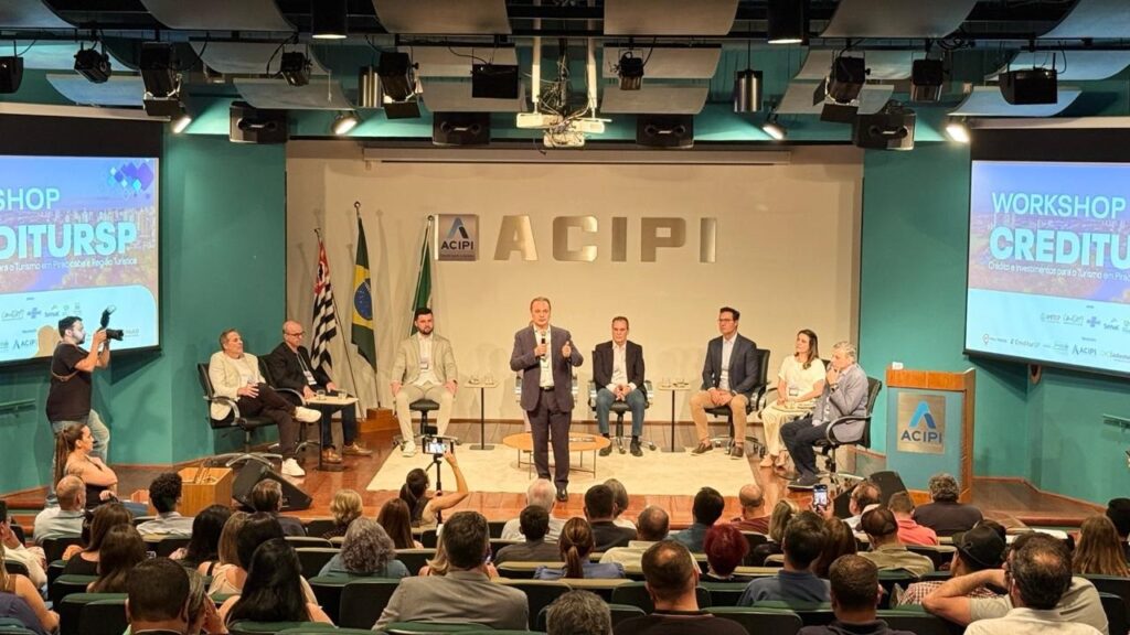 A imagem mostra um evento em um auditório, provavelmente um workshop ou seminário, com palestrantes no palco e uma audiência sentada em cadeiras. No palco, há um grupo de pessoas sentadas e em pé. Um homem de terno escuro está em pé no centro, falando em um microfone, enquanto outras quatro pessoas estão sentadas em cadeiras atrás dele. Há também uma tela grande ao fundo com o título "WORKSHOP CREDITURSP" e "Crédito e Investimentos para o Turismo em Piracicaba e Região Turística". Bandeiras do Brasil e de São Paulo estão visíveis ao lado dos palestrantes. A audiência, composta por muitas pessoas, está sentada em fileiras de cadeiras, voltada para o palco. Alguns membros da audiência estão segurando celulares, possivelmente para tirar fotos ou gravar o evento. Um fotógrafo com uma câmera profissional está posicionado à esquerda do palco, tirando fotos dos palestrantes. A iluminação do auditório sugere que o evento está em andamento.
