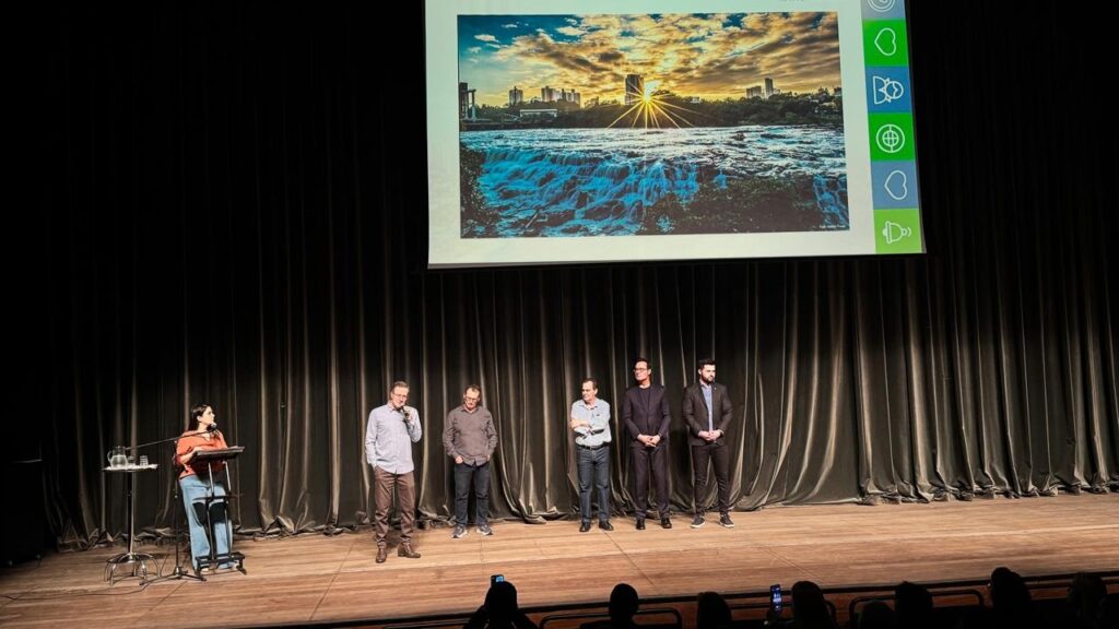 A imagem mostra uma sala de conferências com um palco e uma tela grande ao fundo. No palco, há seis pessoas, sendo uma mulher à esquerda e cinco homens à direita. A mulher está em pé em frente a um púlpito, segurando um microfone. Os cinco homens estão alinhados no lado direito do palco, todos em pé e olhando para a audiência. A tela ao fundo exibe uma imagem de uma paisagem com uma cachoeira e prédios ao fundo, sob um céu dramático ao pôr do sol. À direita da imagem projetada, há uma barra vertical com vários ícones verdes, incluindo um coração, um globo e um símbolo de áudio. O palco é de madeira e as cortinas ao fundo são de cor escura, possivelmente veludo, o que sugere um ambiente formal. Na parte inferior da imagem, vislumbra-se a audiência sentada, com algumas pessoas segurando seus celulares.