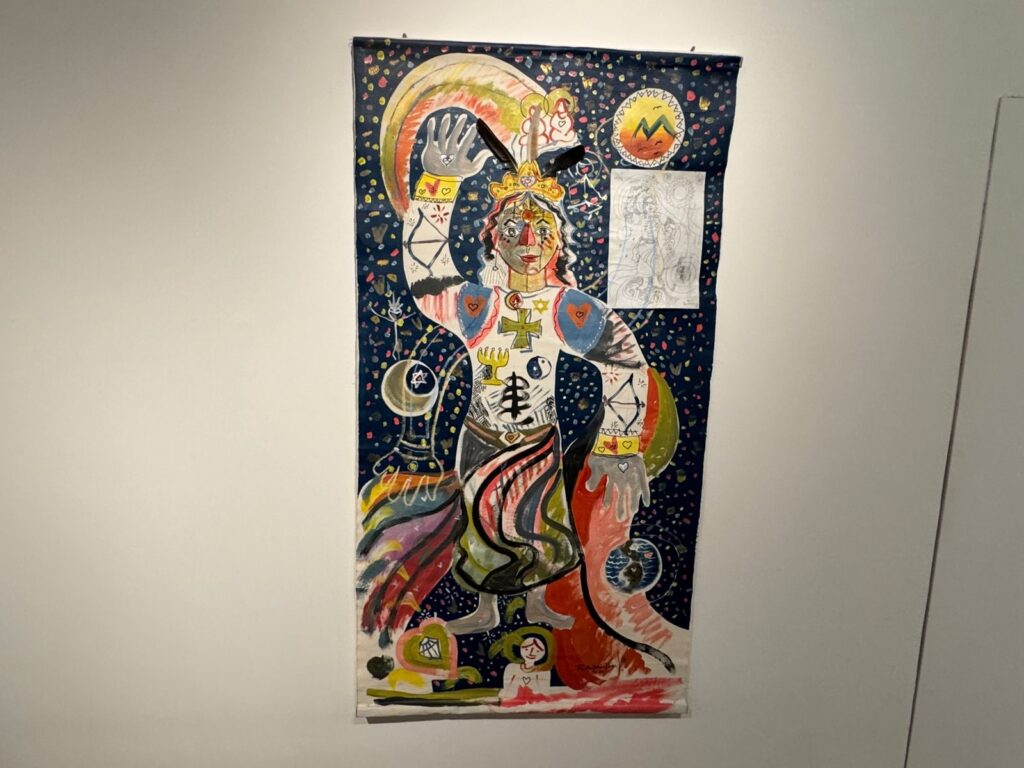 A imagem exibe uma tapeçaria colorida e vibrante, pendurada em uma parede branca. A tapeçaria é dominada por uma figura central estilizada, parecendo uma divindade ou um ser místico. A figura é adornada com uma coroa, joias e vestimentas ricas em detalhes e simbolismos. O fundo da tapeçaria é escuro, pontilhado com pequenas formas que lembram estrelas ou confetes, criando uma atmosfera cósmica. Ao redor da figura central, há vários elementos simbólicos, como luas, estrelas (incluindo uma estrela de Davi), corações e formas geométricas. As cores são intensas e variadas, com predominância de tons de azul, vermelho, amarelo e verde. Em destaque, há duas representações menores afixadas na tapeçaria principal: uma imagem circular à direita superior com uma paisagem montanhosa e um sol estilizado, e abaixo dela, um esboço a lápis de uma figura humana com traços delicados. A tapeçaria parece ser uma obra de arte expressiva e cheia de significado, possivelmente com influências de diversas culturas e crenças.