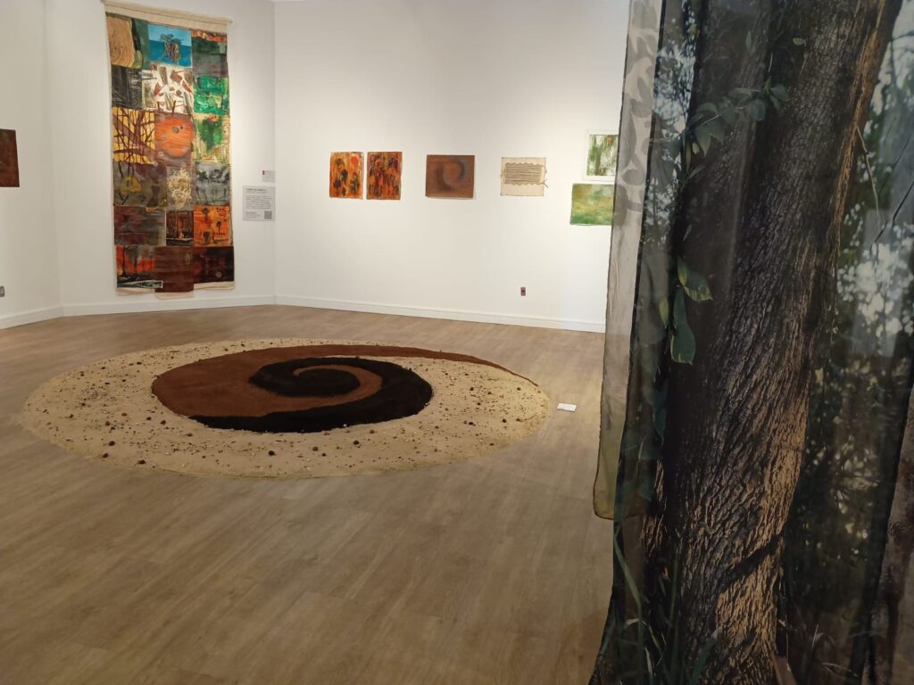 A imagem exibe uma instalação de arte em uma galeria. No centro do espaço, um grande tapete circular em forma de espiral é feito de diferentes tipos de terra e pedrinhas, criando um contraste visual e textural. Ao fundo, pendurado em uma parede branca, há um longo painel têxtil composto por diversos retalhos de tecidos coloridos e texturizados, com padrões abstratos e figurativos. Em outra parede, há uma série de pinturas menores, predominantemente em tons de laranja e marrom, com algumas peças em tons verdes. À direita da imagem, um tecido translúcido com uma estampa de árvore e folhagens cria uma cortina, adicionando profundidade e um elemento natural à composição. O piso da galeria é de madeira clara, complementando o ambiente.