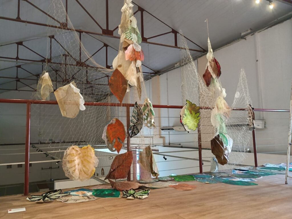 A imagem exibe uma instalação artística suspensa em um espaço interno, possivelmente um museu ou galeria. Elementos que se assemelham a folhas, feitos de materiais diversos como papel, tecido e talvez plástico, estão pendurados por redes finas, lembrando redes de pesca. As "folhas" apresentam uma variedade de cores e texturas, com tons de marrom, laranja, bege, verde e branco, algumas com padrões intrincados ou translúcidas. Elas estão dispostas em diferentes alturas, criando uma sensação de movimento e leveza. A estrutura do edifício, com vigas metálicas aparentes no teto e corrimãos vermelhos, sugere um ambiente industrial ou adaptado para exposições. No chão, há mais elementos decorativos, também em formato de folhas e com representações de água, que complementam a instalação aérea. A iluminação, vinda de cima, realça as formas e cores da obra.