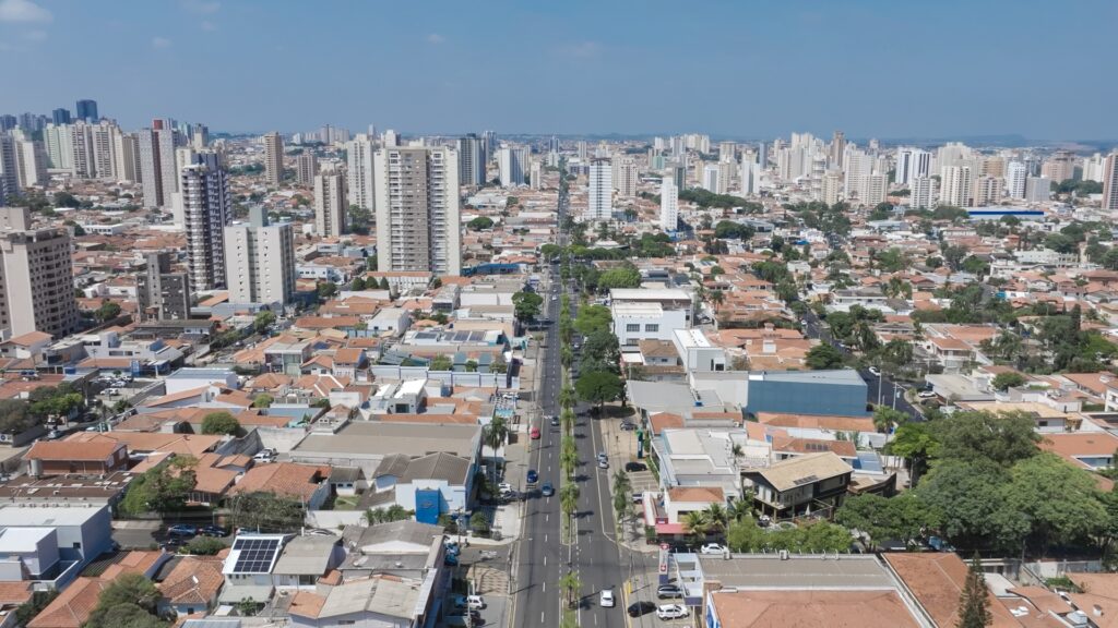 A imagem mostra uma vista aérea de uma cidade grande, com uma longa avenida central que se estende até o horizonte. A avenida é larga, com várias faixas de tráfego, e está ladeada por palmeiras que acompanham todo o seu comprimento. De ambos os lados da avenida, há uma mistura de edifícios residenciais e comerciais. Os edifícios mais próximos são baixos, com telhados vermelhos, enquanto, mais ao fundo, surgem prédios altos com fachadas modernas, muitas vezes de vidro. A cidade parece bastante densa, com muitas construções próximas umas das outras. O céu está claro e azul, indicando um dia ensolarado. A luz do sol cria sombras definidas, evidenciando a textura dos telhados e das árvores. A perspectiva é de um ponto elevado, permitindo uma visão ampla e detalhada da avenida e da cidade ao redor.