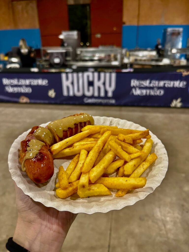 A imagem mostra um prato de papel branco contendo um cachorro-quente com molho e batatas fritas. O cachorro-quente é longo e de cor marrom avermelhada, com fatias transversais que revelam o interior, e é generosamente coberto com um molho amarelado, possivelmente mostarda. Ao lado, há uma porção abundante de batatas fritas douradas, temperadas com especiarias visíveis. Ao fundo, desfocado, observa-se um estande de comida com uma faixa azul que diz "KUCKY Gastronomia" em letras grandes e brancas, ladeada por "Restaurante Alemão" em letras menores. Há também equipamentos de cozinha e pessoas trabalhando no estande, sugerindo que a comida foi adquirida em um evento ou feira gastronômica. A mão que segura o prato está visível na parte inferior esquerda da imagem.